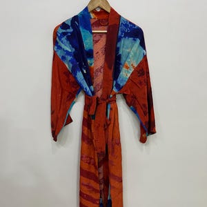 Puede incluir: Una bata estampada vibrante en tonos de rojo, naranja y azul cuelga de una percha de madera. La bata presenta un diseño de cintura anudada y un patrón abstracto colorido. Las mangas son largas y la tela parece ligera.