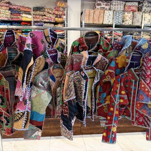 May include: A collection of colorful, patchwork jackets hanging on a clothing rack. The jackets feature a variety of patterns and colors, including floral, geometric, and abstract designs. The jackets are made of various fabrics and have a quilted appearance.