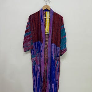 May include: A long, open-front kimono robe featuring a vibrant tie-dye pattern in shades of deep blue, magenta, and teal. The garment has three-quarter length sleeves, two front pockets, and a contrasting yellow and white tie-dye neck label.