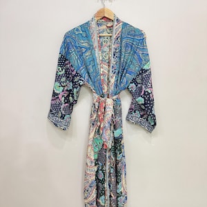 May include: A long-sleeved robe featuring a patchwork design with various blue, black, and multi-colored floral and paisley patterns. The garment includes a matching fabric tie belt at the waist and a shawl collar, constructed from lightweight fabric.