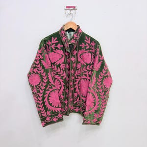 Könnte beinhalten: Eine grüne Samtjacke mit aufwendiger rosa Blumenstickerei. Die Jacke hängt an einem Holzkleiderbügel vor einer weißen Wand. Die Stickerei zeigt detaillierte Blumen- und Blattmuster, die einen auffälligen Kontrast zum dunkelgrünen Stoff bilden.