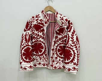 May include: A white jacket with red floral embroidery. The jacket is hanging on a wooden hanger against a white wall. The interior lining features red and white vertical stripes.