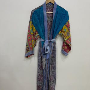 May include: A colorful, patterned robe with a tie belt, hanging on a wooden hanger. The robe features a mix of blue, yellow, and pink floral and geometric patterns. The sleeves and body have different designs, creating a patchwork effect.