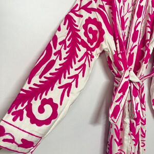 Puede incluir: Batista blanca con un patr&oacute;n de bordado floral rosa. La bata tiene mangas largas y un cintur&oacute;n de lazo.