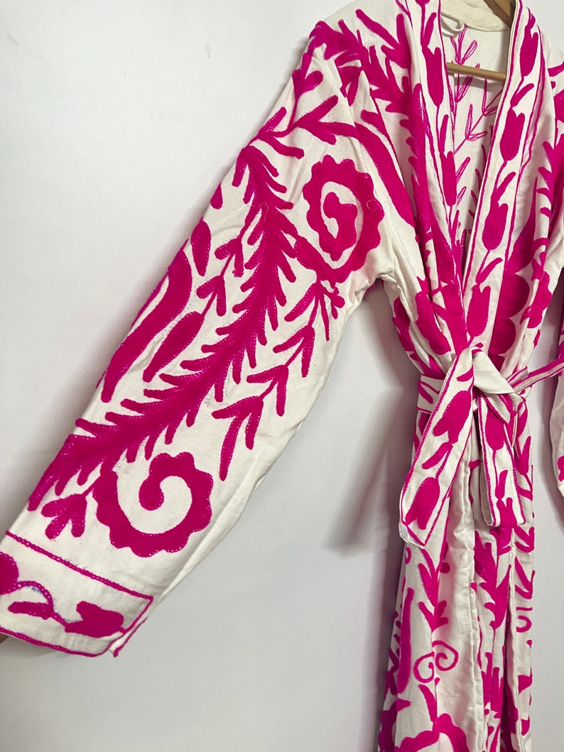 Puede incluir: Batista blanca con un patr&oacute;n de bordado floral rosa. La bata tiene mangas largas y un cintur&oacute;n de lazo.