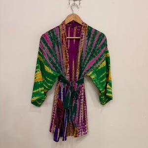 May include: A short kimono robe featuring a vibrant tie-dye pattern in shades of purple, green, and yellow. The garment has wide sleeves, an open front, and a matching fabric tie belt at the waist, all hanging from a wooden hanger.