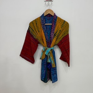 May include: A colorful, patterned robe with a tie belt, hanging on a wooden hanger. The robe features a mix of colors, including red, yellow, blue, and purple. The tie belt is a mix of blue and gold. The robe is displayed against a white wall.