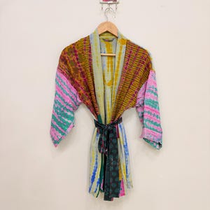 Może przedstawiać: Lekki szlafrok typu kimono z patchworkowym wzorem i motywami tie-dye w kolorach musztardowym, różowym, morskim i białym. Ubiór posiada długie rękawy, otwarty przód oraz pasujący pasek z tkaniny w ciemny kwiatowy wzór.