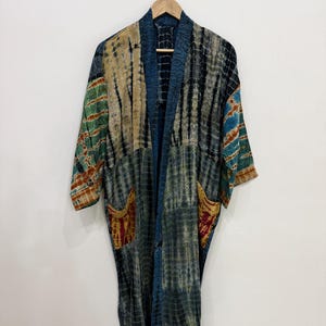 Puede incluir: Una bata estilo kimono de manga larga con un diseño de patchwork y patrones tie-dye en tonos azul, verde, amarillo y marrón. La prenda cuenta con un cuello azul oscuro sólido, mangas anchas y dos bolsillos delanteros con detalles en rojo.