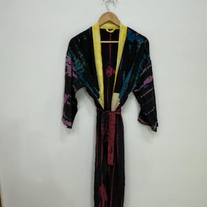 May include: A long-sleeved robe featuring a black base with tie-dye patterns in shades of purple, blue, and pink. The garment includes a bright yellow collar and a matching fabric tie belt at the waist, hanging from a wooden hanger.