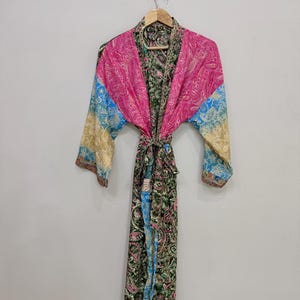 May include: A long-sleeved robe featuring a patchwork design with vibrant pink, blue, and green paisley patterns. The garment includes a matching fabric tie belt at the waist and is displayed on a wooden hanger against a plain wall.