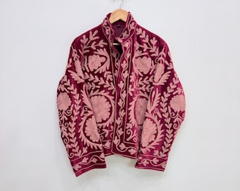 Puede incluir: Chaqueta de terciopelo burdeos con intrincados bordados florales en un tono rosa más claro. La chaqueta, con cuello alto, está colgada de una percha de madera. El bordado cubre el frente, las mangas y la espalda de la chaqueta.