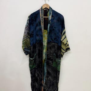 Può includere: Un lungo kimono aperto sul davanti, nei toni del blu, nero, verde e giallo. L'indumento presenta un motivo tie-dye e due tasche anteriori. È appeso a una gruccia di legno contro un muro bianco.