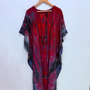 May include: A red, black, and blue tie-dye caftan dress with a V-neck and short sleeves. The dress is hanging on a wooden hanger. The fabric has vertical stripes of red, black, and blue.