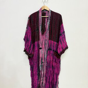 Può includere: Un lungo kimono aperto sul davanti caratterizzato da un vivace motivo tie-dye nelle tonalità del magenta, bordeaux scuro e nero. Il capo presenta maniche ampie e larghe, un collo a scialle e una silhouette fluida che cade dritta dalle spalle.