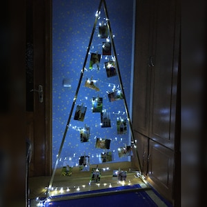Könnte beinhalten: Ein dreieckiger Fotorahmen aus Holz, verziert mit Lichterketten. Kleine Fotos sind mit Wäscheklammern am Rahmen befestigt. Die Präsentation steht vor einer blauen Wand mit Sternenmuster und schafft eine warme Atmosphäre.