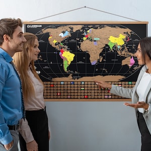 May include: A large world map with a black background and gold scratch-off surface is displayed on a wall. The map is framed in wood and has country outlines in various colors. The map is labeled "LANDMASS WORLD". A group of people are looking at the map.