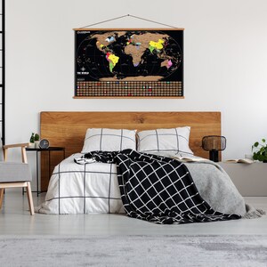 May include: A black world map with gold foil scratch-off areas hangs above a bed. The map is framed with a wooden hanger. The bedroom features a wooden headboard, a patterned chair, and a black and white patterned blanket.
