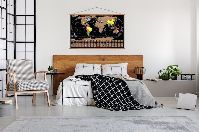 May include: A black world map with gold foil scratch-off areas hangs above a bed. The map is framed with a wooden hanger. The bedroom features a wooden headboard, a patterned chair, and a black and white patterned blanket.
