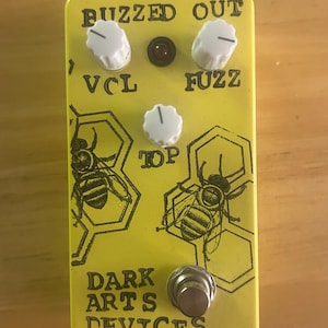 May include: Yellow guitar effects pedal with the text "BUZZED OUT", "VCL", "FUZZ", and "TOP". The pedal has white knobs, a red light, and a silver footswitch. The design includes bee illustrations and the text "DARK ARTS DEVICES".