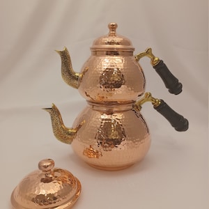 May include: A two-tiered copper teapot with hammered texture and black wooden handles. The teapot has a gold-colored spout and lid.