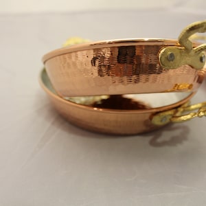 Op de afbeelding: Twee koperen kookpotten met gehammerde oppervlakken en goudkleurige handvatten. De potten zijn op elkaar gestapeld.