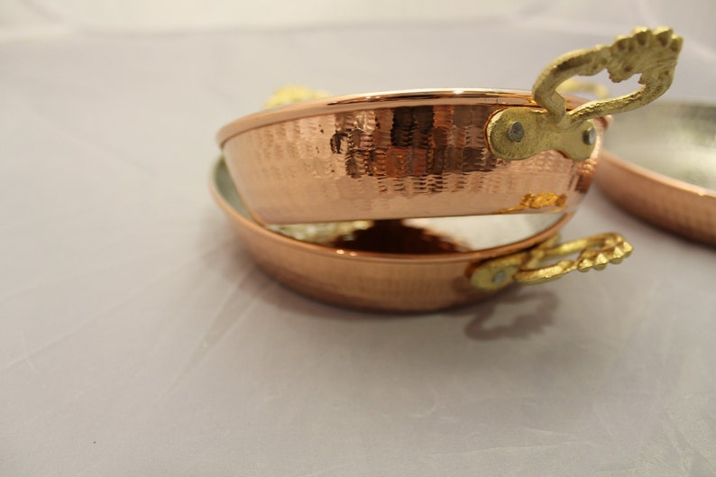 Op de afbeelding: Twee koperen kookpotten met gehammerde oppervlakken en goudkleurige handvatten. De potten zijn op elkaar gestapeld.