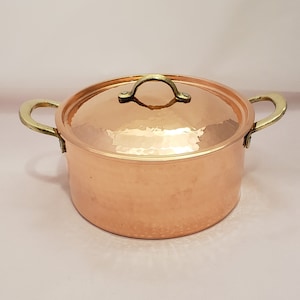 May include: A copper pot with a hammered finish and brass handles. The pot has a lid with a brass handle.
