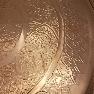 May include: A round copper tray with an intricate embossed design. The design features a repeating pattern of stylized flowers and leaves.