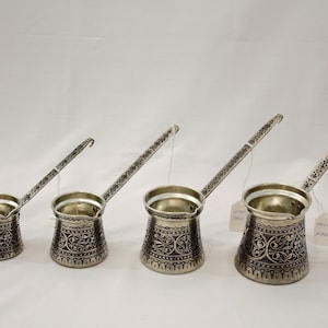 Może przedstawiać: Cztery srebrne tureckie kawiarki z ozdobnymi wzorami i długimi uchwytami. Kawiarki są ułożone według rozmiaru, od najmniejszej po lewej stronie do największej po prawej.