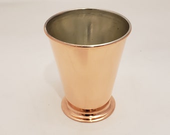 May include: A copper-colored metal cup with a flared rim and a rounded base. The cup has a shiny, polished finish.
