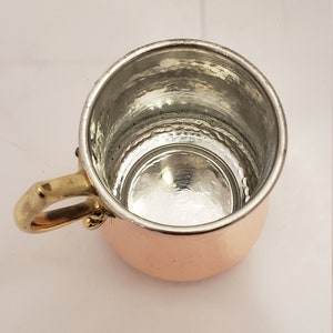 Pode incluir: Uma pequena caneca de cobre com acabamento martelado e uma al&ccedil;a de lat&atilde;o. A caneca est&aacute; vazia e tem um interior brilhante e reflexivo.