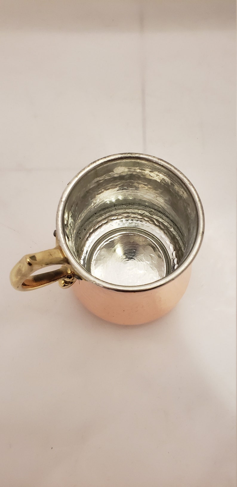 Pode incluir: Uma pequena caneca de cobre com acabamento martelado e uma al&ccedil;a de lat&atilde;o. A caneca est&aacute; vazia e tem um interior brilhante e reflexivo.