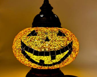 May include: A decorative Halloween lamp shaped like a jack-o'-lantern. The lamp features a mosaic design with yellow and orange glass pieces, a black base, and a black top with a decorative finial. The jack-o'-lantern face is made of black tiles.