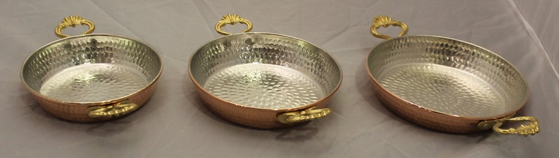 Op de afbeelding: Drie ovale koperen serveerschalen met gehammerde oppervlakken en goudkleurige handvatten.