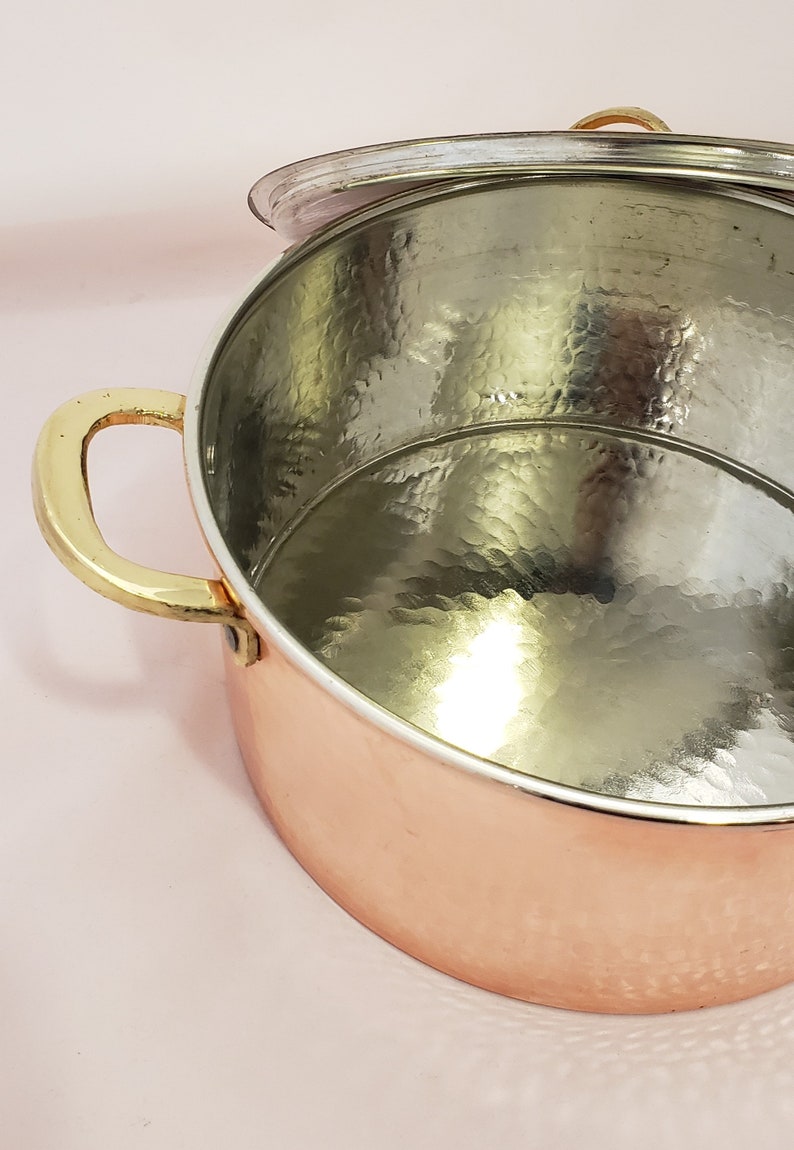 May include: A copper pot with a hammered finish and a brass handle. The pot has a lid that is slightly ajar. The pot is sitting on a pink surface.