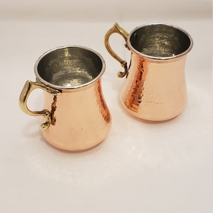 Pode incluir: Duas canecas de cobre martelado com al&ccedil;as de lat&atilde;o. As canecas t&ecirc;m um acabamento met&aacute;lico brilhante e s&atilde;o perfeitas para servir bebidas.