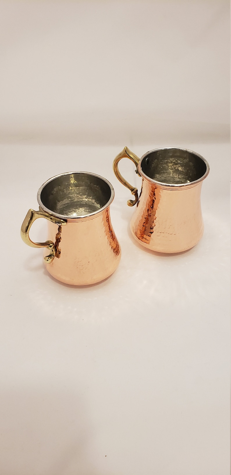 Pode incluir: Duas canecas de cobre martelado com al&ccedil;as de lat&atilde;o. As canecas t&ecirc;m um acabamento met&aacute;lico brilhante e s&atilde;o perfeitas para servir bebidas.