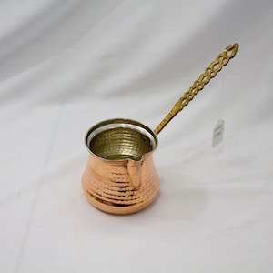 May include: A copper coffee pot with a hammered finish and a brass handle. The pot has a small spout and a rounded body.