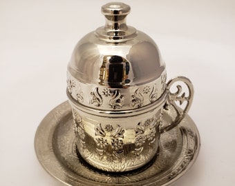 May include: A silver Turkish coffee set with intricate floral designs. The set includes a small pot with a lid and a matching saucer.