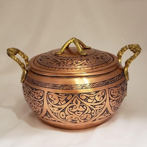 May include: A copper-colored, lidded bowl with ornate, etched patterns. The bowl has two gold-colored handles and a gold-colored handle on the lid. The design features intricate floral and geometric motifs, creating a decorative and functional piece.