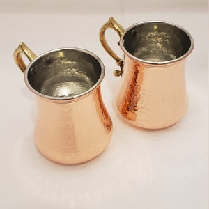 Pode incluir: Duas canecas de cobre martelado com al&ccedil;as de lat&atilde;o. As canecas t&ecirc;m um acabamento de cobre brilhante e s&atilde;o perfeitas para servir bebidas.