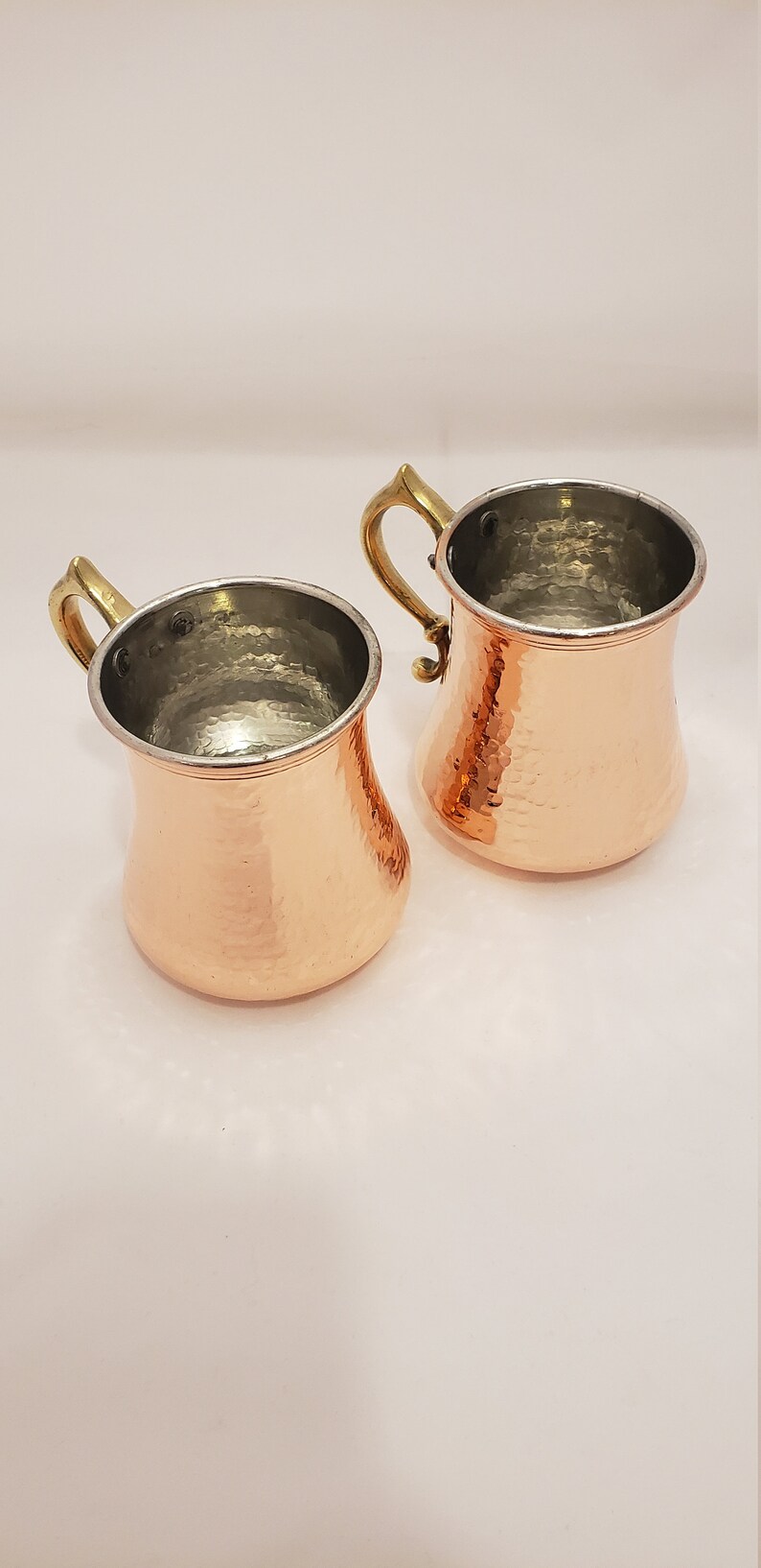 Pode incluir: Duas canecas de cobre martelado com al&ccedil;as de lat&atilde;o. As canecas t&ecirc;m um acabamento de cobre brilhante e s&atilde;o perfeitas para servir bebidas.