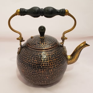 May include: A hammered copper teapot with a black wooden handle and a gold spout. The teapot has a textured surface and a round body.
