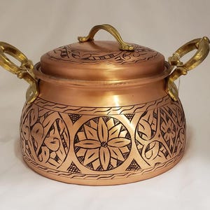 May include: A copper cooking pot with a lid and two brass handles. The pot features intricate etched floral and geometric patterns around the body. The lid also has etched designs and a brass handle.