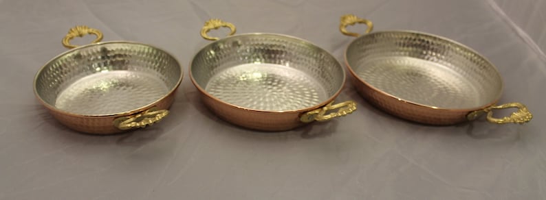 Op de afbeelding: Drie koperen serveerschalen met gehammerde oppervlakken en goudkleurige handvatten. De schalen zijn in elkaar genest, met de grootste schaal achterin en de kleinste schaal vooraan.