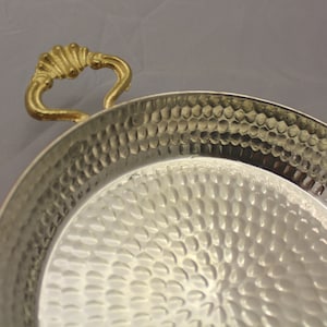 Op de afbeelding: Een gehamerd koperen serveerschaal met een gouden handvat. De schaal is rond en heeft een getextureerd oppervlak.