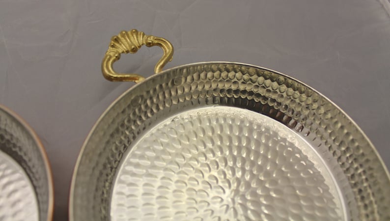 Op de afbeelding: Een gehamerd koperen serveerschaal met een gouden handvat. De schaal is rond en heeft een getextureerd oppervlak.