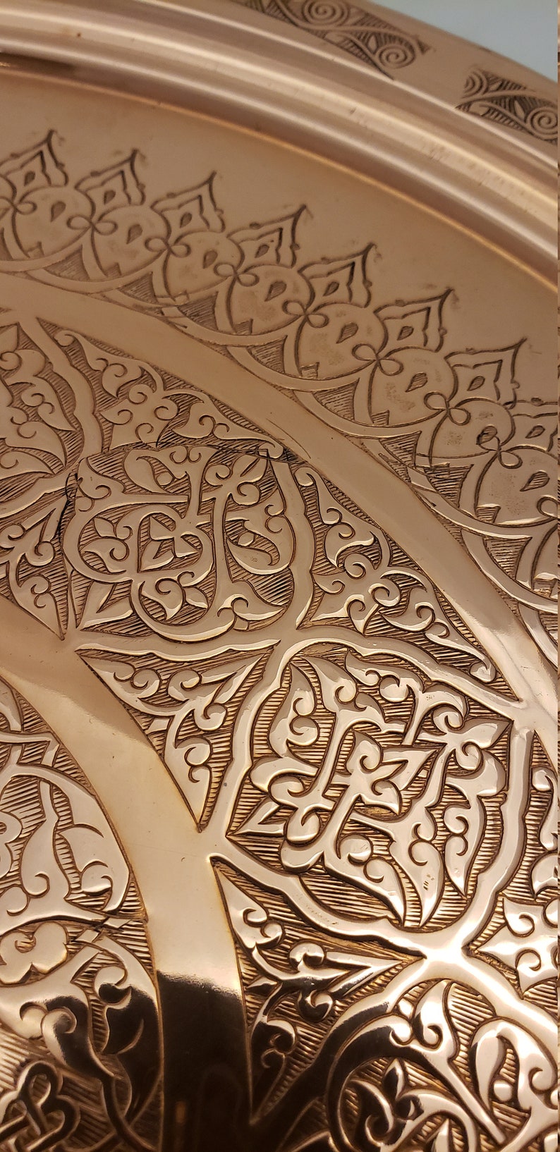 May include: Close-up of a copper tray with intricate embossed floral patterns. The tray has a warm, metallic sheen and a slightly rounded shape.