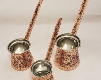 May include: Three copper Turkish coffee pots with ornate engraved designs. The pots have long handles and are arranged on a white surface.